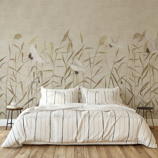 A neatly made bed with cream bedding and striped pillows, placed in front of a large wall mural showing reeds and cranes in beige and brown tones. Two small side tables and floor lamps sit on either side, with a light wood floor below.