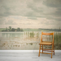 Beverley Wallpaper with a wooden chair in front of a large wall mural of a landscape with water and trees.