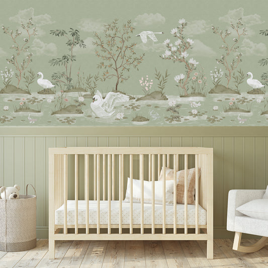A nursery room scene showing a light wood crib with white bedding, a woven basket with a plush toy, and a white rocking chair against a wall decorated with a soft sage green and beige toile-style wallpaper featuring swans, trees, and water lilies.