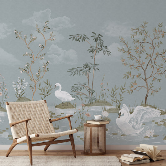 A styled interior scene showing a large wall mural with a soft blue-gray background and a delicate botanical watercolor design featuring swans, reeds, and trees. In front of the mural is a woven rattan armchair, a small round side table with books and a cup, and a few open books on the floor.
