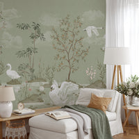 A styled living room featuring a sage green wallpaper with a delicate landscape design of swans, trees, and clouds. The room is furnished with a white armchair, a matching ottoman, a white floor lamp, and a side table with flowers.