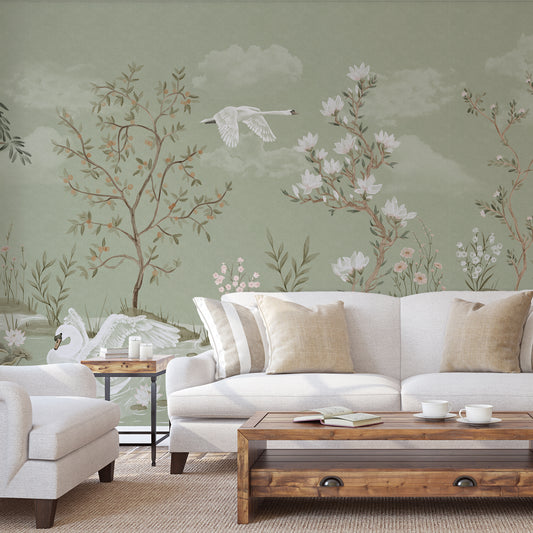 A styled living room featuring a sage green wall mural with delicate botanical illustrations of trees, flowers, and white swans, behind a white sofa with beige pillows and a wooden coffee table.