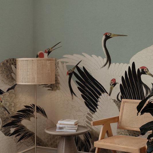 A styled interior corner featuring a large wall mural with detailed cranes in black, white, and gray tones against a muted sage green wall. A woven rattan floor lamp stands on the left, a small round table with stacked books sits in the center, and a wooden chair with a woven back is on the right.