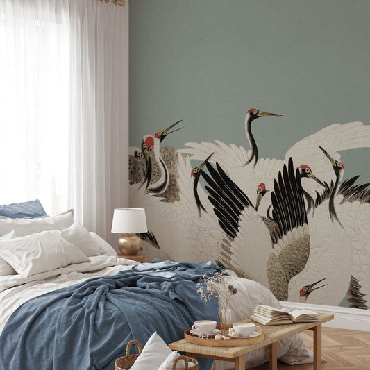 A styled bedroom interior featuring a large wall mural with black, white, and gray cranes on a muted sage green background. The room includes a bed with white and blue bedding, a small wooden table with cups and books, sheer white curtains, and a gold floor lamp.