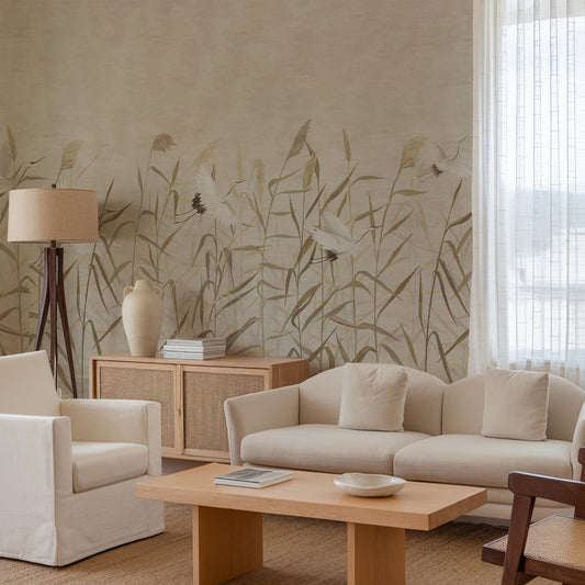 A styled living room featuring a large wall mural with a delicate botanical pattern of leaves and birds in muted beige and brown tones. The room is furnished with a cream-colored sofa, matching armchair, light wood coffee table, a wooden sideboard with a beige ceramic vase, and a floor lamp.