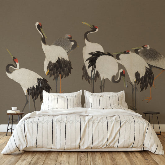 A styled bedroom scene with a bed dressed in white bedding featuring thin black vertical stripes, set against a large wall mural of cranes on a taupe wall. Two small side tables sit on either side of the bed.