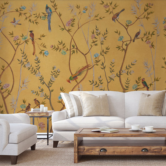 A styled living room featuring a prominent mustard yellow wallpaper with a detailed botanical pattern of branches, leaves, and colorful birds. In front of the wall is a white fabric sofa with neutral beige pillows, a wooden coffee table, and a matching white armchair.
