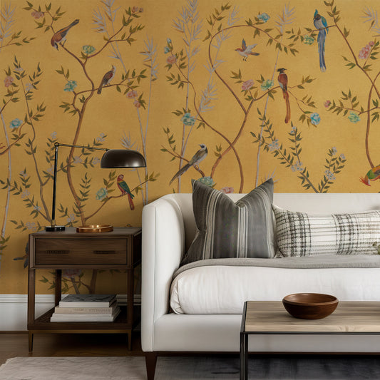 A styled living room corner featuring a mustard yellow wallpaper with a detailed botanical and bird pattern, styled with a white sofa, striped and plaid pillows, a dark wood side table with a black lamp, and a coffee table.